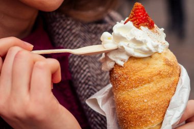 İştahlı bir kadın Prag caddesinde eko ahşap kaşık kullanarak vanilyalı ve çilekli geleneksel Çek tatlısı Trdelnik yiyor.