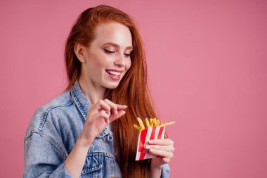 Şirin, uzun, seksi, kızıl saçlı kadın pembe stüdyoda patates kızartması ve karton kutu tutuyor.