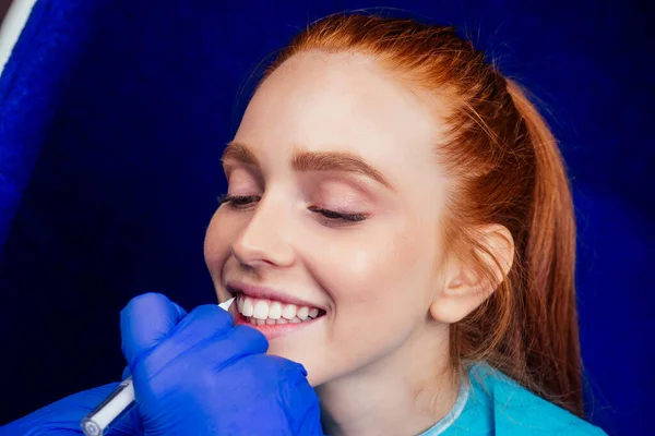 Dentist works on tooth crown in dental laboratory ultraviolet painting teeth with mineral and calcium cream to young red-haired ginger woman — Stock Photo, Image