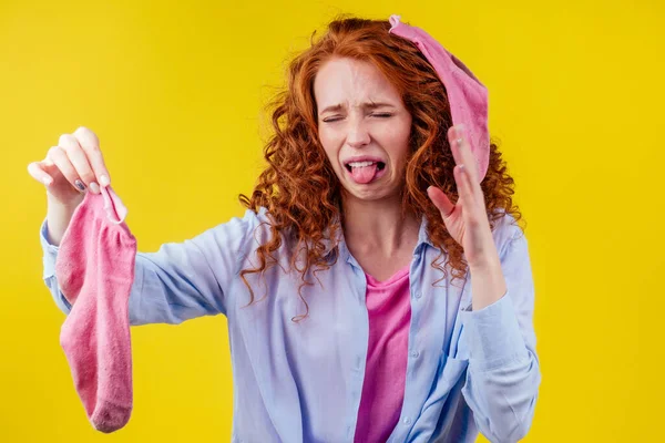 Кудрявая рыжая женщина в хлопчатобумажной рубашке жест плохо пахнет держа грязный розовый носок из отвратительный взгляд на ее лице в студии желтый фон — стоковое фото