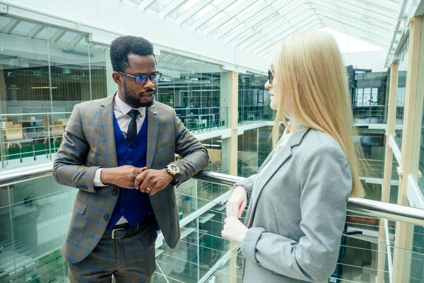 Homme afro-américain dans une chemise blanche en veste brune posant parler à une femme caucasienne blonde d'affaires dans un costume gris dans un bureau moderne avec des fenêtres en verre centre commercial — Photo