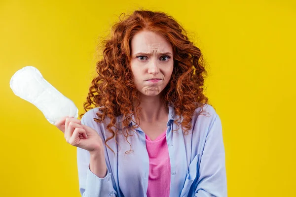 Embaraço ruiva ruiva encaracolado mulher olhando timidez e segurando almofadas sanitárias em estúdio fundo amarelo Imagem De Stock