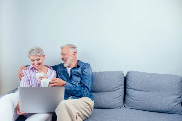 Пожилая пара в любви сидя серый диван в гостиной посмотрите на экран ноутбука и пить чашку чая или кофе подражания — стоковое фото