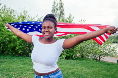 Gülümseyen Afro-Amerikalı kadın elinde ABD bayrağı ve sonbahar akşamı gölün kenarındaki parkta kameraya bakıyor.