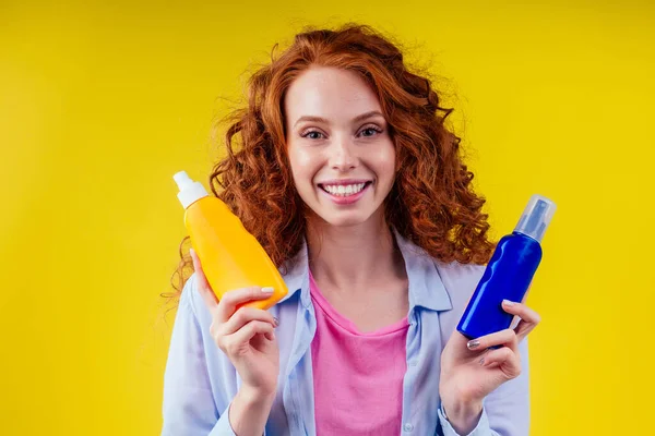 Kızıl saçlı kıvırcık saçlı kadın elinde güneş kremi şişesiyle stüdyo sarı arka planda doğru seçimi yapıyor. — Stok fotoğraf