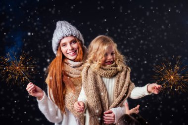 redhaired ginger woman in warm hat and little blonde girl wear knitted scarf holding bengal sparkler in studio black background clipart