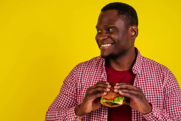 黄色の背景に隔離されたハンバーガーを食べる若いアフリカ系アメリカ人男性 — ストック写真
