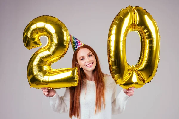 Förvånad och förvånad radhaired ingefära kvinna med födelsedagshatt horn håller ballonger 20 års jubileum siffra i studio vit bakgrund — Stockfoto