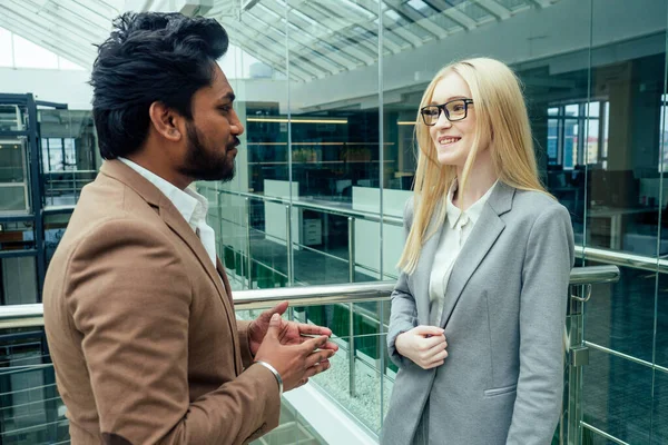 Indian man in a white shirt in brown jacket posing talking to a business blonde caucasus woman in a gray suit in a modern office with glass windows shopping mall — Stock Photo, Image