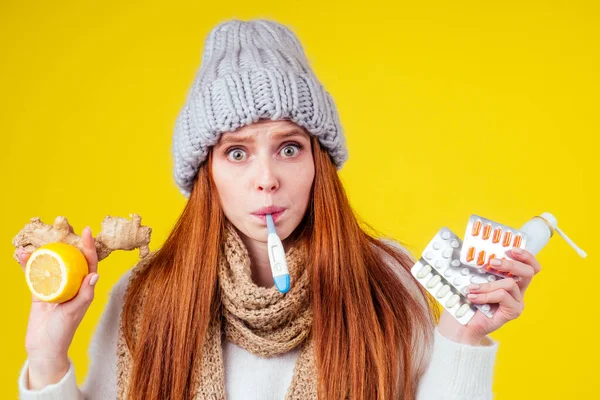 Enfermedad enferma mujer pelirroja con suéter de punto, sombrero con bufanda elegir entre limón natural y jengibre remedio popular para los resfriados y montón de píldoras en fondo amarillo estudio — Foto de Stock