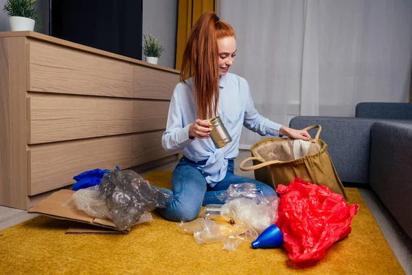 Felice ragazza rossa zenzero raccolta rifiuti: bottiglia, carta e plastica seduta a tappeto pavimento in soggiorno in un appartamento confortevole e moderno — Foto Stock