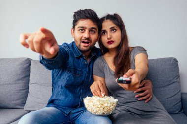 Şaşırmış genç Hintli çift ellerinde patlamış mısırla ellerinde uzaktan kumandayla gerilim filmi ya da futbol izliyorlar.