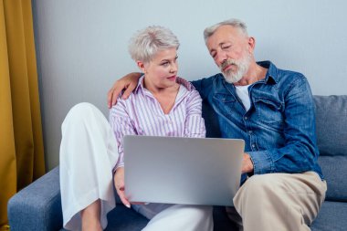 Oturma odasında gri koltukta oturan aşık yaşlı çift ekran laptopuna bakıyor.
