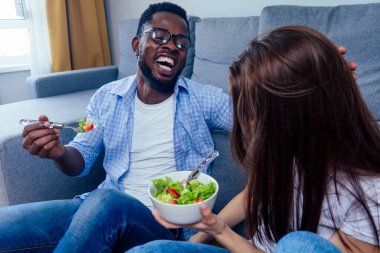 Afro-Amerikalı bir adam oturma odasında kadınla salata yiyor.