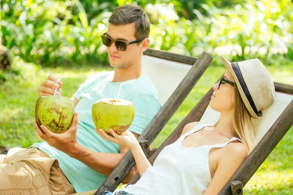 Молодая любящая пара пьет кокос, сидя под пальмами в шезлонге курорта — стоковое фото