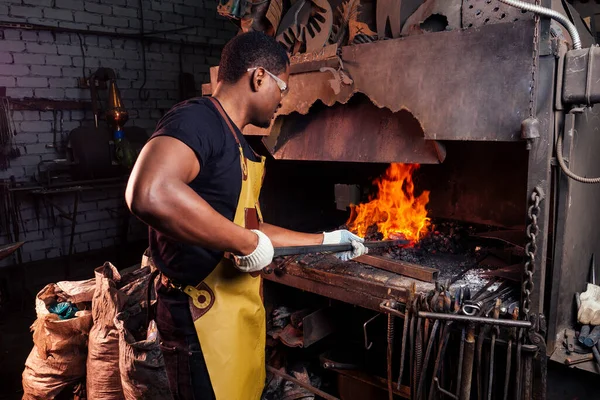 Een knappe Afrikaans-Amerikaanse man die staal smeed naast een oven in een donkere werkplaats. kleine ondernemingen, — Stockfoto