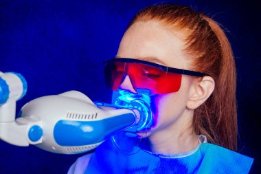 Kızıl saçlı, kızıl saçlı, kızıl saçlı, koruyucu kırmızı gözlüklü, dişçi ofisinde oturan ve yumurta sandalyesinde gülümseyen genç, neşeli bir bayan müşteri.