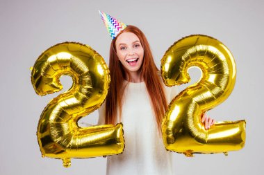 Şaşırmış ve şaşkın kızıl saçlı kadın elinde balonlar tutuyordu. Stüdyonun beyaz arka planında 22. yıl dönümü numarasıydı.
