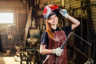 Strong and worthy pretty redhead ginher woman wearing protection helmet and leather apron with gloves holding welding machine instrument . small business concept clipart