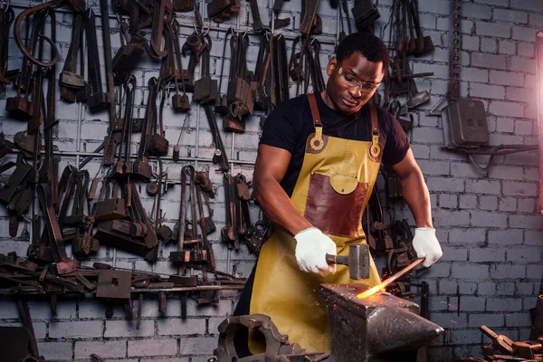 Martillo industria pequeña empresa concept.african hombre americano vestido con ropa histórica está martillando en el yunque. Un herrero forja un producto metálico —  Fotos de Stock