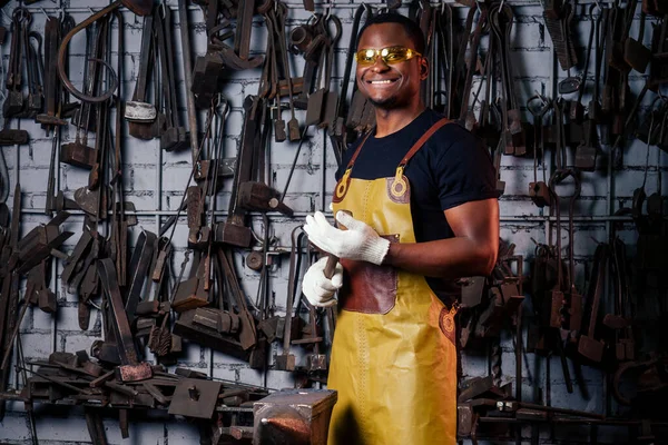 Jóképű afro-amerikai kovács férfi munkás dolgozik műhelyben, visel bőr kötény és védőszemüveg kesztyű. — Stock Fotó