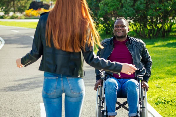 Hombre afroamericano sentado en silla de ruedas, su novia pelirroja rodando cochecito en el parque de otoño. tener una cita romántica — Foto de Stock