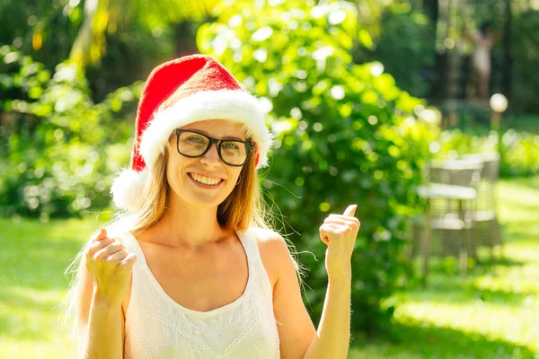 Närbild av en ung flicka i glasögon och en röd hatt av jultomten på jul i nyår på bakgrund sommar havet palm i trädgården resort — Stockfoto
