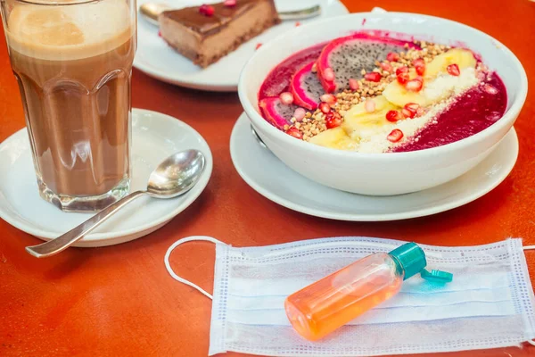 woman useing antibacterial gel on hands before start to eating her healthy food in tropical asian cafe . smoothie bowl , raw cake and coffee on table with sanitizer on bottle