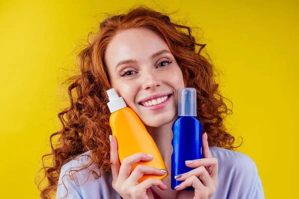 Kızıl saçlı kıvırcık saçlı kadın elinde güneş kremi şişesiyle stüdyo sarı arka planda doğru seçimi yapıyor. — Stok fotoğraf