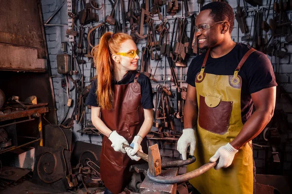Pár v lásce zrzavý zázvor mladá evropská žena a afro-americký muž nosí koženou zástěru pracovní kovář workshop.small rodina mezinárodní obchodní koncept — Stock fotografie