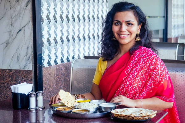 Krásná brunetka žena v červeném sari jíst s apetitem tradiční thali wirh vzestup, tvaroh, dal v restauraci Goa masala čaj — Stock fotografie