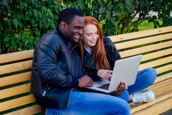 Internationell utbildning koncept.mörkhyade man wuth rödhårig ingefära kvinna tittar på laptop och sitter på bänk, gröna löv bakgrund — Stockfoto