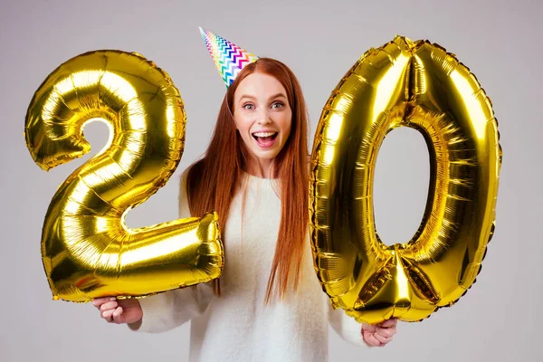 Förvånad och förvånad radhaired ingefära kvinna med födelsedagshatt horn håller ballonger 20 års jubileum siffra i studio vit bakgrund — Stockfoto