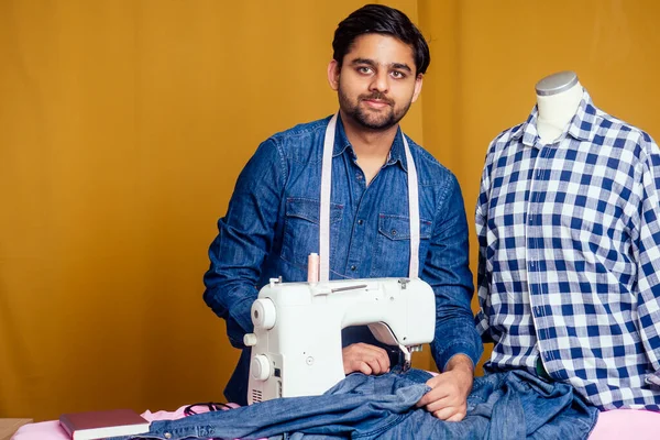 handsome indian tailor man In a stylish shirt working with dreen jeans cotton textile at home workshop yellow curtain background