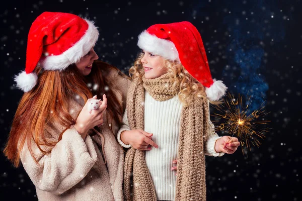 redhaired ginger woman in warm sheeps wool coat and santa claus hat and little blonde girl wear knitted scarf holding bengal sparkler and white rat black background snow in dark night