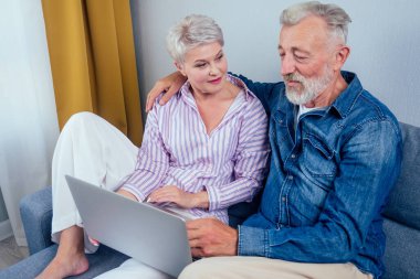 Oturma odasında gri koltukta oturan aşık yaşlı çift ekran laptopuna bakıyor.