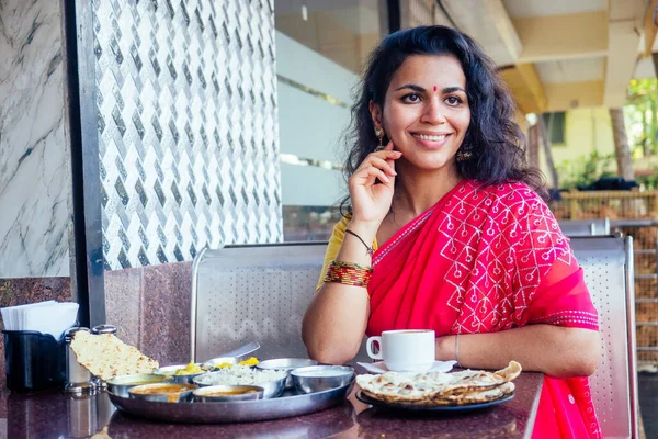 Hermosa morena en sari rojo comiendo con apetito tradicional thali con subida, cuajada, dal en el restaurante Goa y bebiendo té masala —  Fotos de Stock