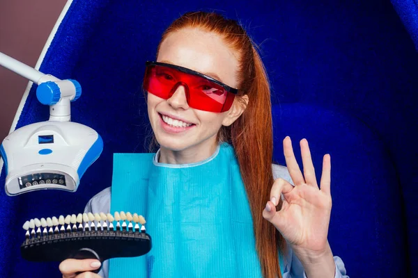 Mujer pelirroja pelirroja feliz usando vidrio rojo en los ojos, mostrando un gesto bien y sosteniendo la paleta del espectro dental junto al aparato ultravioleta blanqueador en la silla de huevo — Foto de Stock