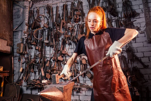 Roodharige roodharige vrouw die smithytools gebruikt in donkere werkplaatsen om hamer hamer details te smeden. concept voor kleine ondernemingen — Stockfoto