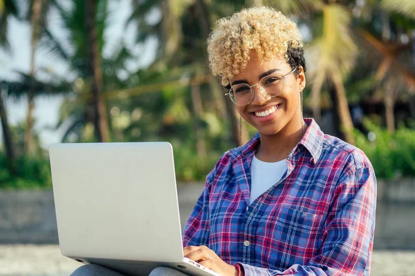 Glad latinska frilansande kvinna med blonda afro lockar sitter på tom strand med laptop palmer bakgrund. fjärrdröm bästa arbetskoncept — Stockfoto