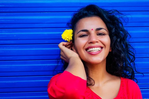 Привлекательная красивая счастливая молодая латиноамериканка с тиковым бинди на лбу, улыбающаяся на фоне синей стены улицы, держа в руках свежий фловер — стоковое фото
