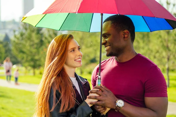 Pelirroja mujer pelirroja y hombre afroamericano de pie cerca el uno del otro bajo el paraguas en el parque de verano — Foto de Stock