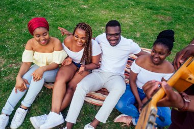 Gülümseyen mutlu Afro-Amerikalı arkadaşlar kampanyanın açık hava pikniğinde gitar çalıyorlar.