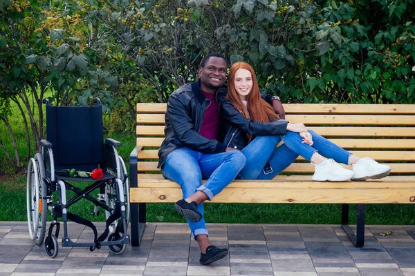 Концепція страхування подорожей. міжнародна пара закоханих сидить на лавці в парку поруч з колісним кріслом — стокове фото