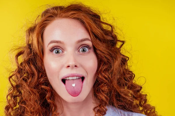 Ruiva ruivo afro encaracolado mulher pele perfeita tomando pílulas de amor feliz no estúdio amarelo fundo .heart forma pílula na língua feminina — Fotografia de Stock