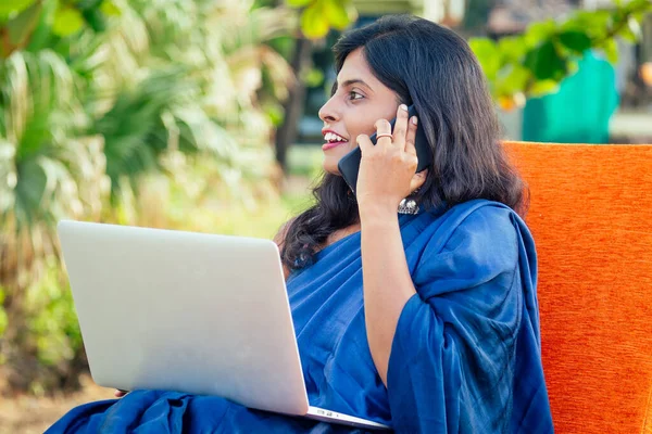 Деловая женщина из Индии азиатская фрилансер, сидящая в кафе. женщина в индийском синем стильном сари сари работает на ноутбуке и улыбается, разговаривая на смартфоне. работа мечты в райском побережье — стоковое фото
