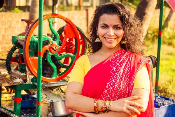Hermosa mujer indígena en sari rojo de pie cerca de la máquina de aparatos de fabricación de jugo de caña de azúcar, plantación de fondo de la granja de verano. pequeña empresa puesta en marcha —  Fotos de Stock
