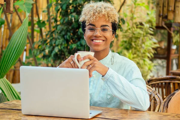 Afrikaans Amerikaans zakenmeisje met schattig krullend blond haar met behulp van laptop en hebben een kopje koffie pauze in de zomer tropische tuin cafe — Stockfoto