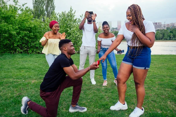 American male standing on one knee asking woman marriage giving ring proposing surprised shocked while gathering group friends all around in shock confusing evening cloudy autumn spring in the park — Stock Photo, Image