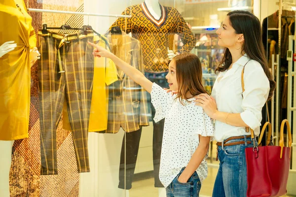 Retrato de uma jovem mulher indiana vestindo roupa moderna t-shirt branca e jeans com sua filha bonito escolher um novo vestido no shopping, vitrine de vidro com manequins — Fotografia de Stock